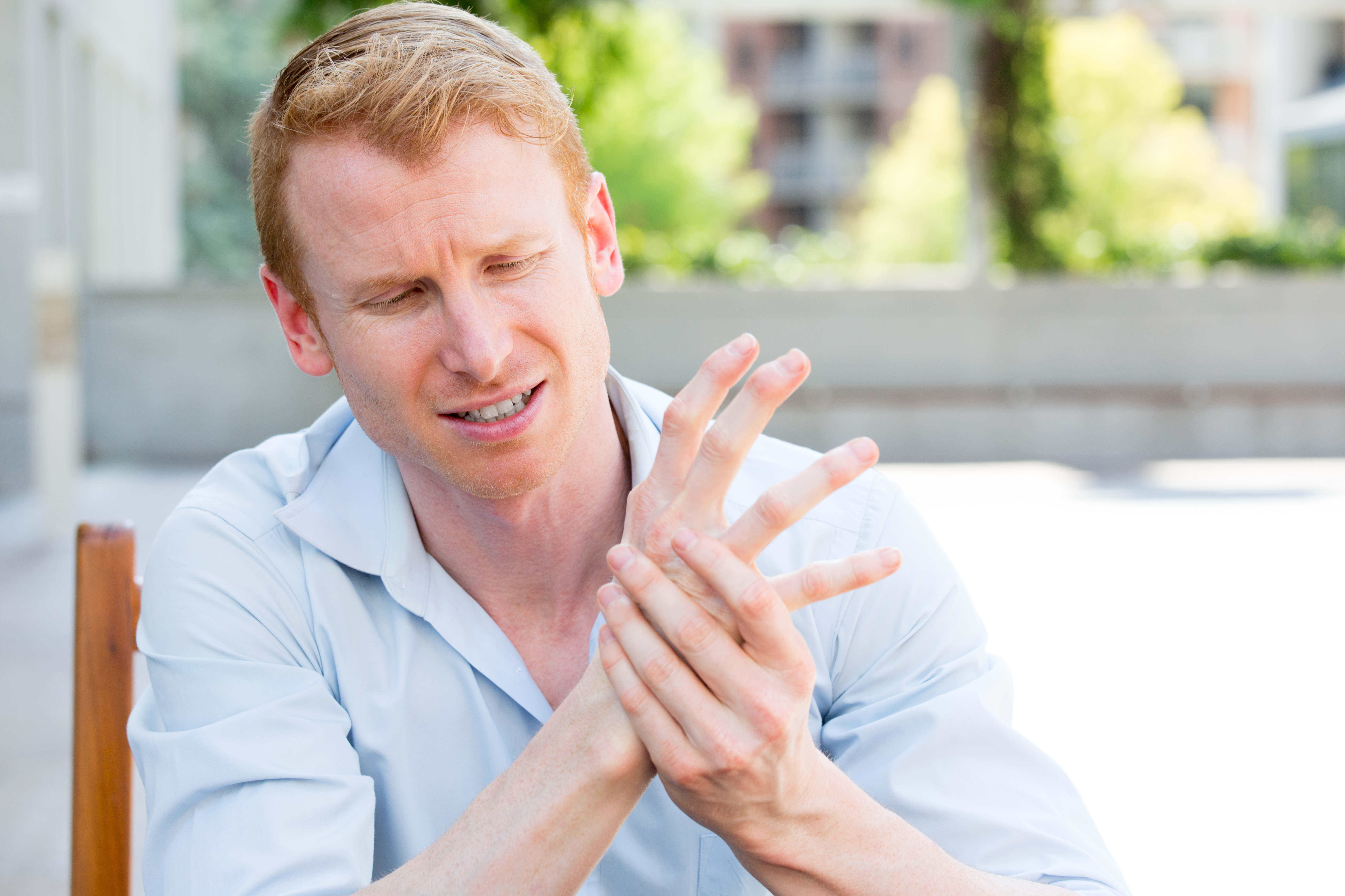 Closeup portrait, young man having acute bad joint pain in his hands, wrtier's cramp, massaging them, sitting in chair, isolated outdoors background