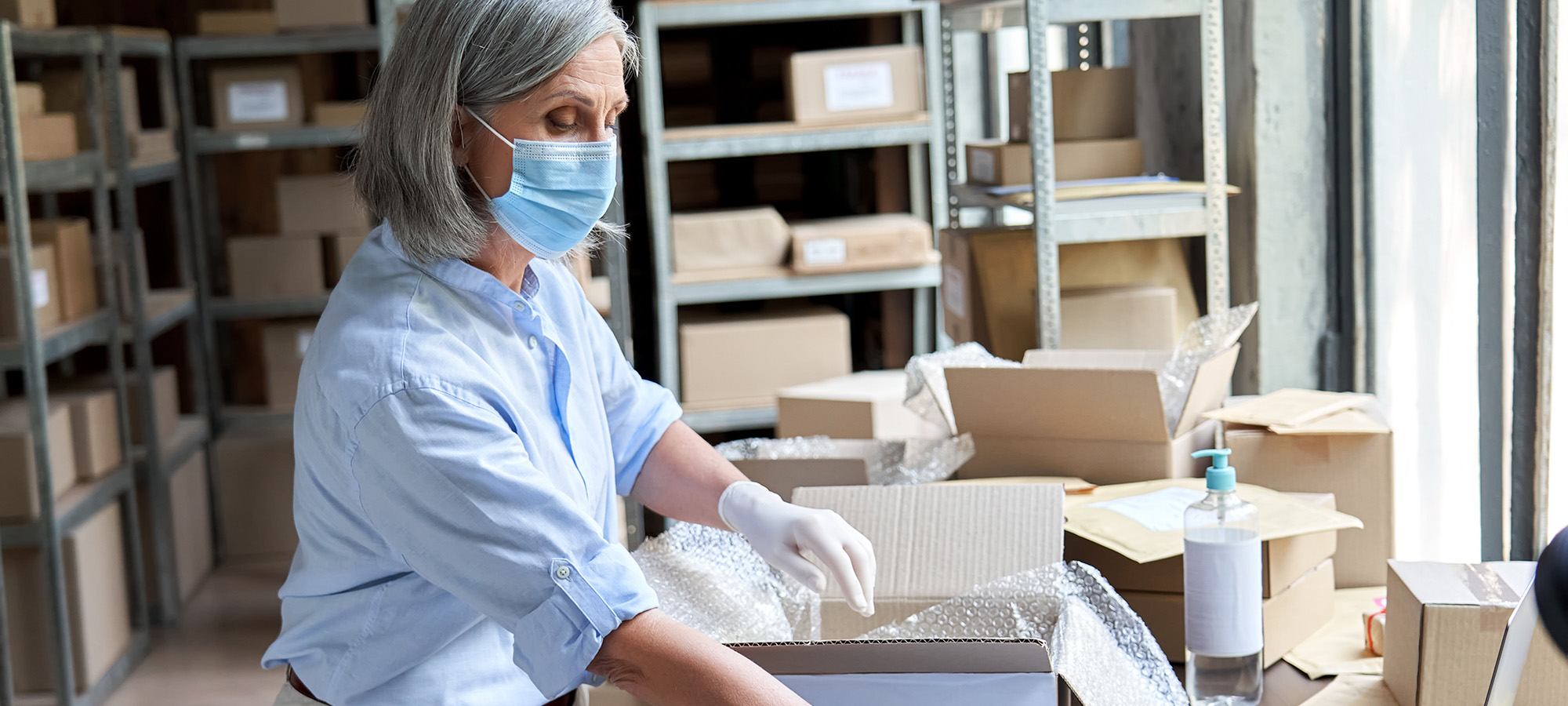 Mature female volunteer warehouse worker wearing face mask working in shipping delivery charitable stock organization packing medical donations box. Covid 19 coronavirus donating and volunteering.