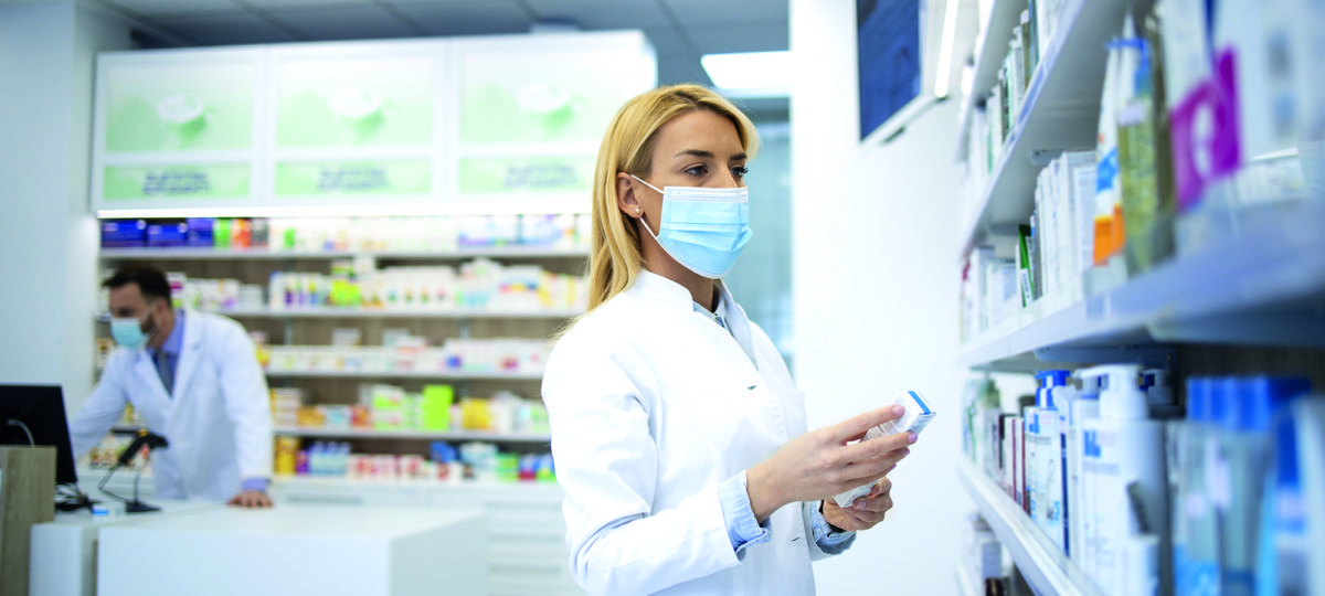 Female pharmacist wearing face mask and white coat holding medicine in pharmacy store during corona virus pandemic.