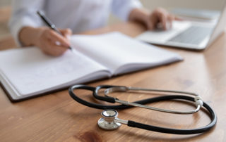 Female doctor physician sits at workplace using laptop, writing notes in medical journal. Woman therapist general practitioner wearing white coat working at table, close up view. Focus on stethoscope