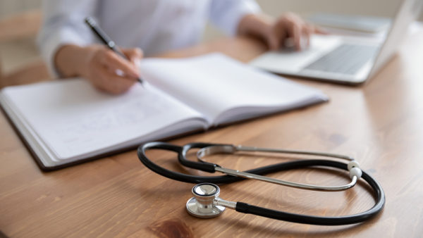 Female doctor physician sits at workplace using laptop, writing notes in medical journal. Woman therapist general practitioner wearing white coat working at table, close up view. Focus on stethoscope
